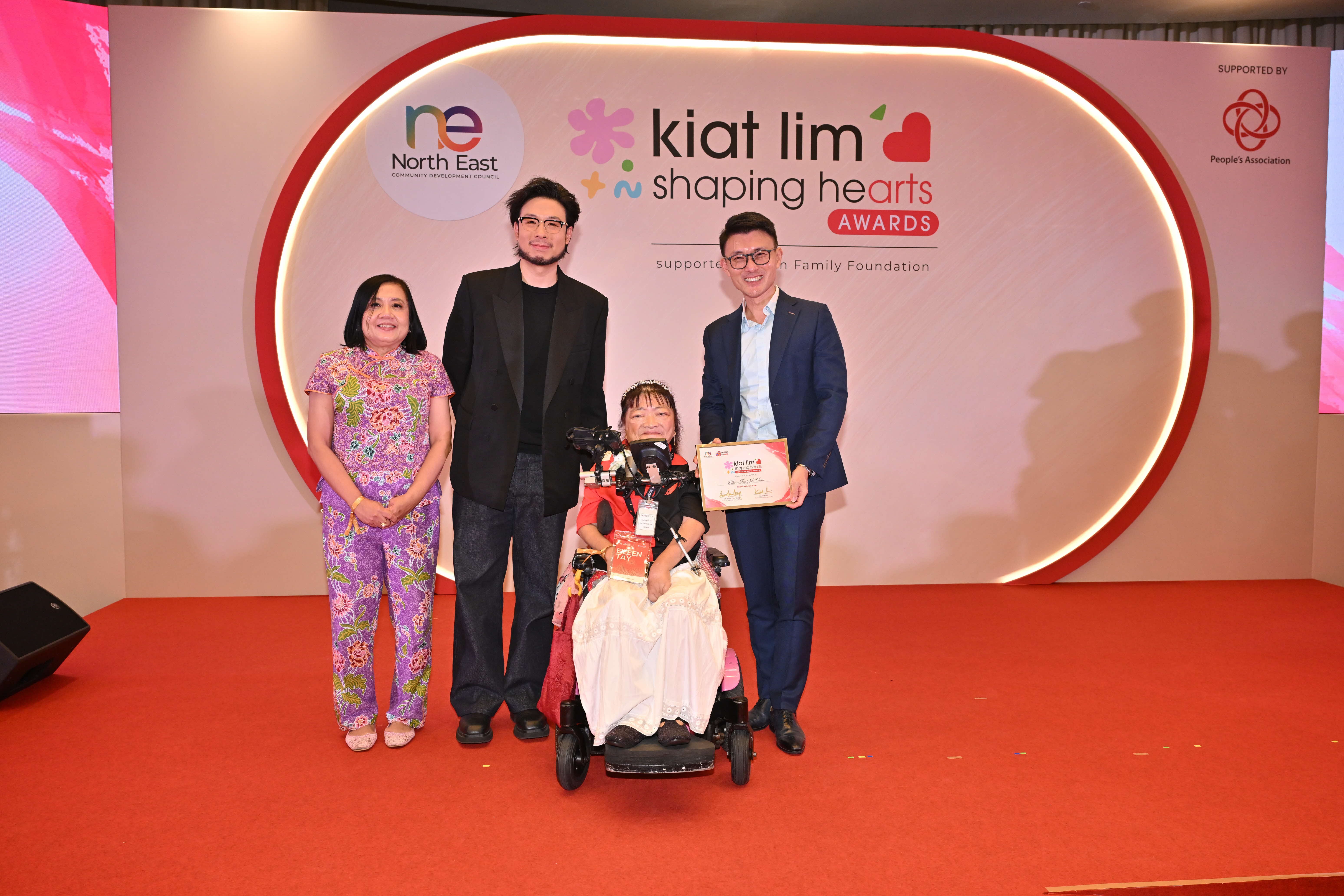 Award recipient standing on stage with the presenter, holding their certificate and posing for a group photo during the award ceremony, with a backdrop reading ‘Kiat Lim-Shaping Hearts Awards'.